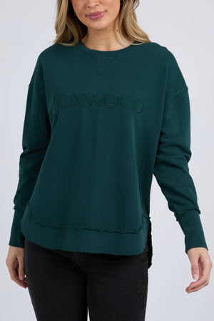 A woman wears the Foxwood Simplified Crew, a dark green crew neck sweatshirt with Foxwood embroidery on the front, paired with black jeans and standing against a plain light background.
