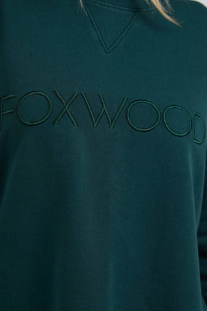Close-up of the Foxwood Simplified Crew, a dark green 100% cotton sweatshirt with "FOXWOOD" embroidered in matching green across the chest.