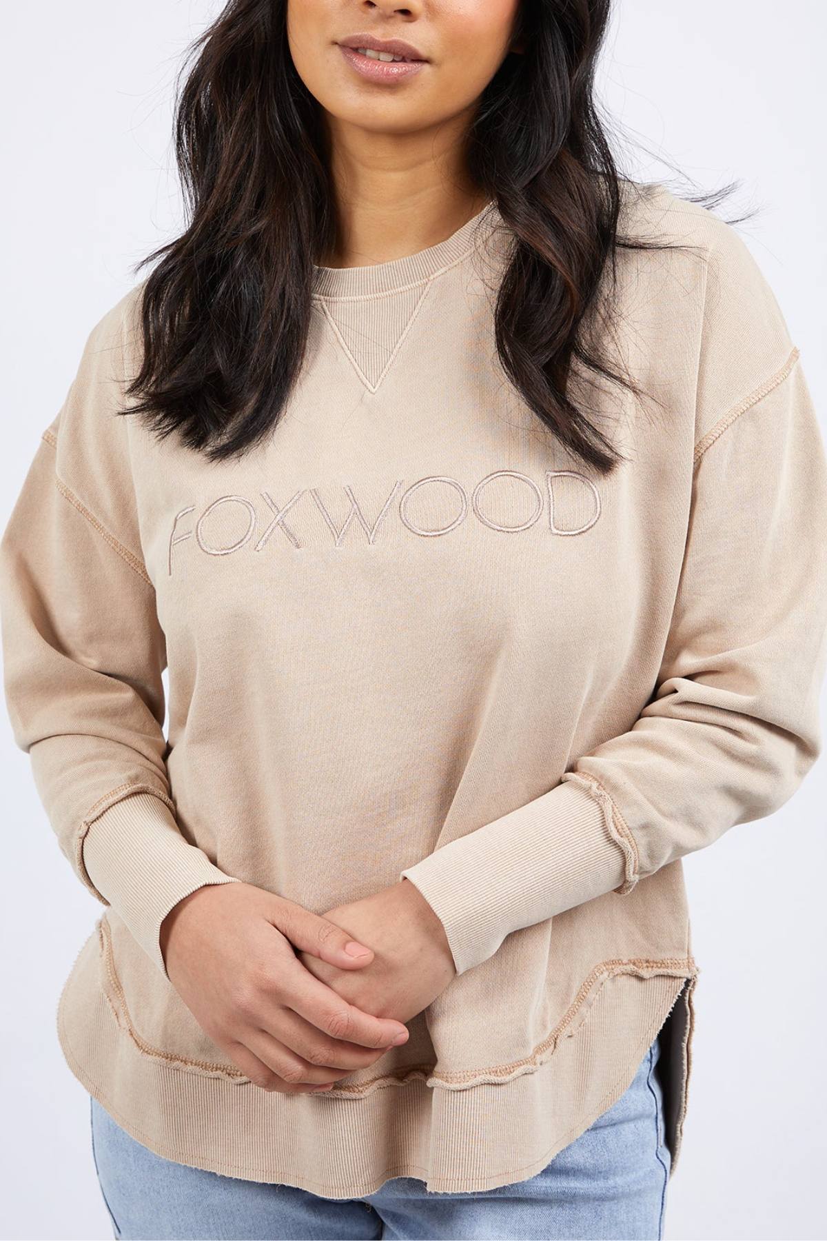A person wears a beige Foxwood Simplified Crew with frayed edges and Foxwood embroidery, paired with light blue jeans and long dark hair, standing against a plain light background.
