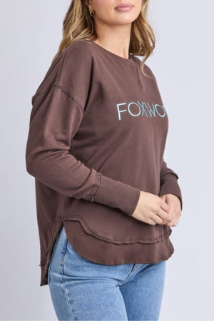 A woman in a loose-fitting brown Foxwood Simplified Metallic Crew sweatshirt with a light blue logo and light blue jeans stands against a plain light gray background.