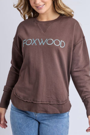 A woman with wavy, lurex-threaded hair smiles while wearing the Foxwood Simplified Metallic Crew sweatshirt in brown with a light blue Foxwood logo across the chest, paired with light blue jeans.