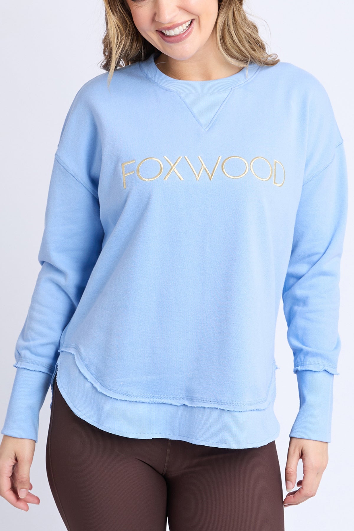 A woman wearing the Foxwood Simplified Metallic Crew, a women's crew sweatshirt with "FOXWOOD" in gold lurex thread, paired with brown leggings, stands against a plain light background and smiles.
