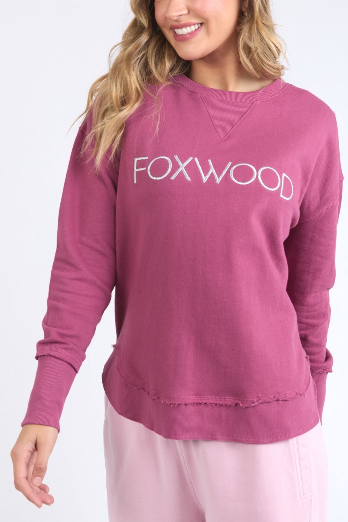 A woman with wavy blonde hair wears the Foxwood Simplified Metallic Crew, featuring Foxwood embroidery on the front. Made from 100% cotton, she pairs it with light pink pants and smiles against a plain white background.