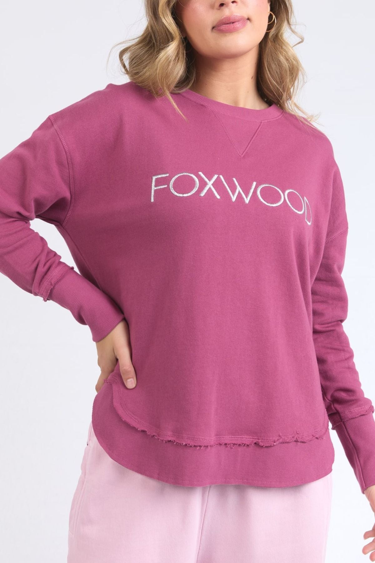 A woman wears the Foxwood Simplified Metallic Crew, a 100% cotton mauve sweatshirt with metallic Foxwood embroidery, paired with light pink pants. She has wavy, shoulder-length hair and poses with one hand on her hip.