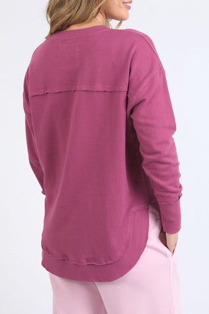 A woman with loose brown hair stands with her back to the camera, wearing a mauve Foxwood Simplified Metallic Crew (100% cotton) and light pink pants. Her right hand is in her pocket. The background is plain white.