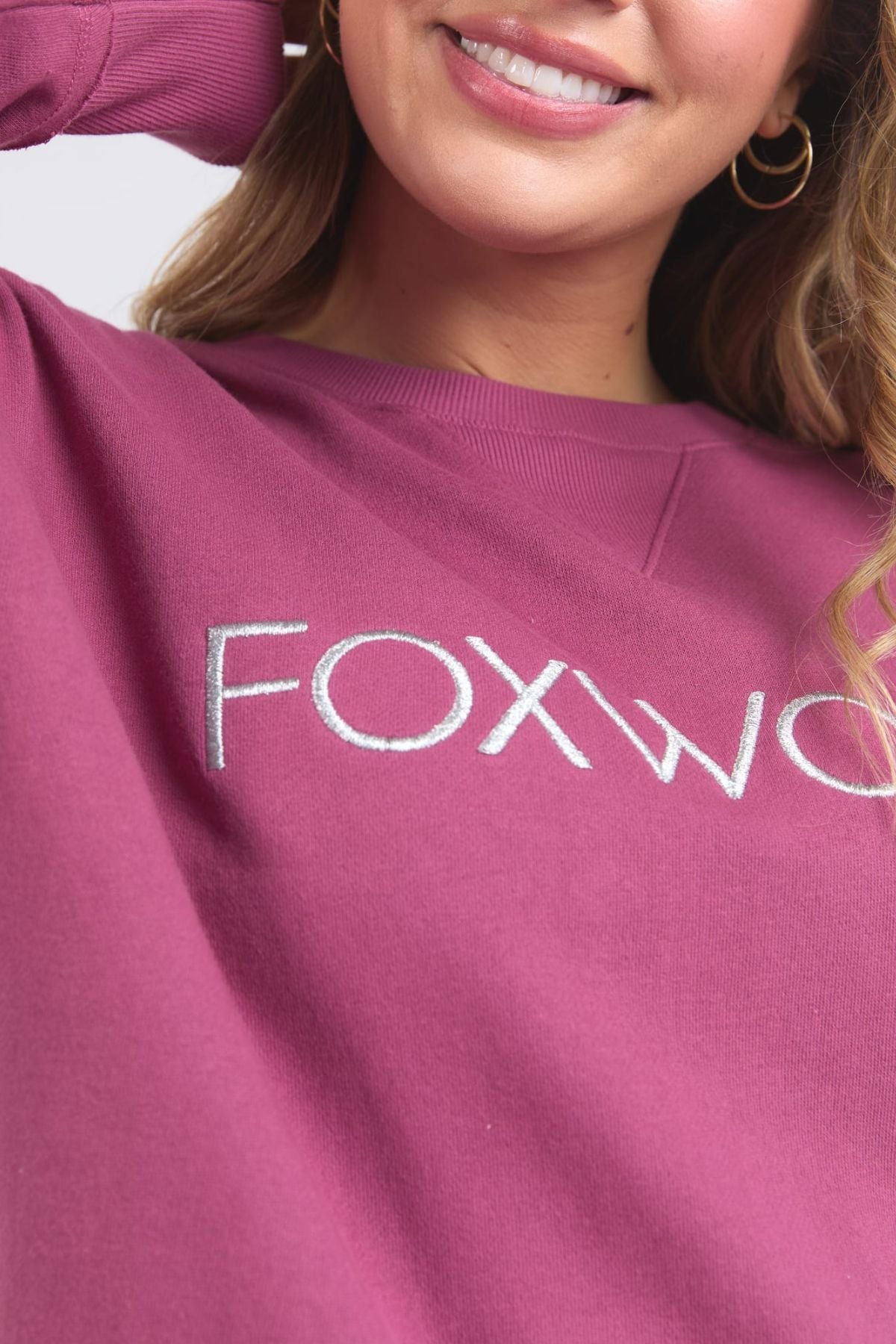 A woman with wavy blonde hair and gold hoop earrings smiles while wearing the Foxwood Simplified Metallic Crew, a magenta 100% cotton sweatshirt with bold metallic Foxwood embroidery; only her lower face and upper torso are visible.