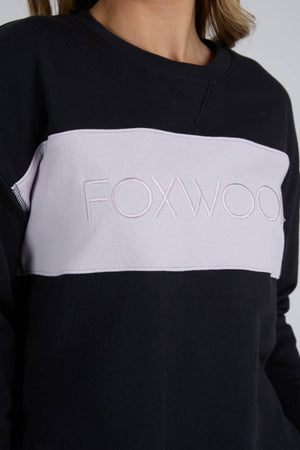 Wearing the Foxwood Simplified Tonal Crew, a black cotton fleece sweater with a light lavender chest panel, bold "FOXWOOD" embroidery, and a chic hi-lo hemline.