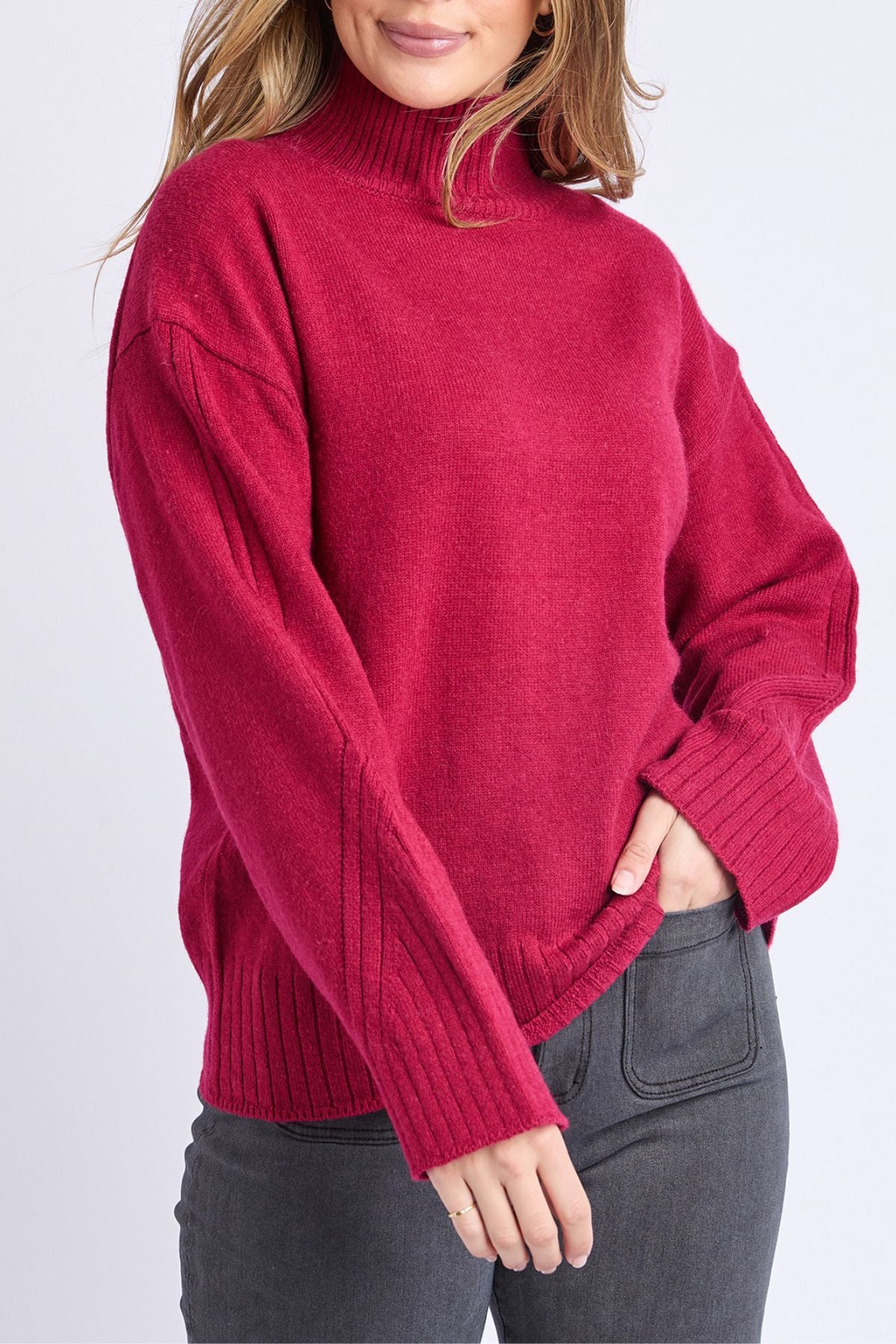Wearing the Foxwood Stellar Knit, a long-sleeved dark pink turtleneck sweater, and dark gray jeans, a person stands with one hand in their pocket against a light background, their face partly out of view.