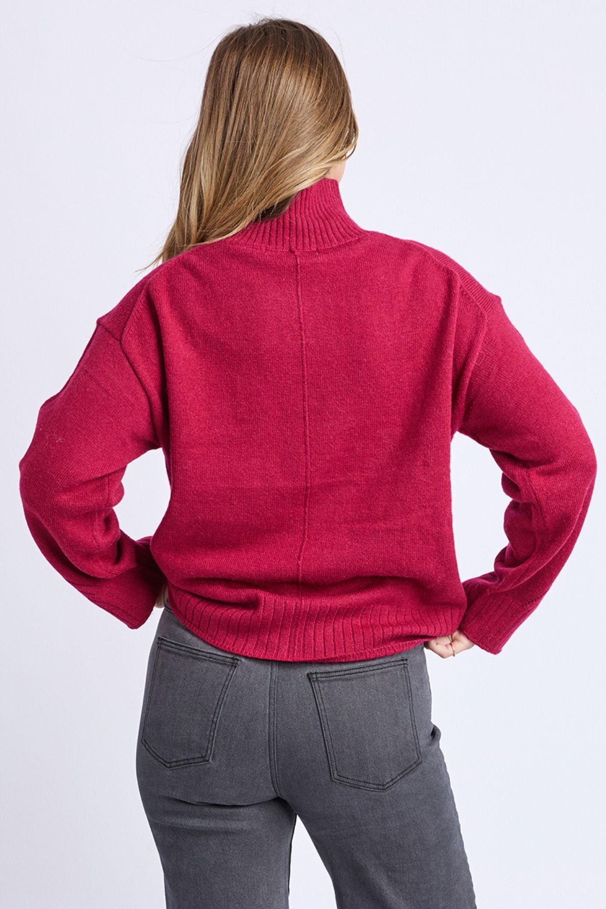 A person with long light brown hair is seen from the back, wearing the Foxwood Stellar Knit red turtleneck and dark gray jeans, standing against a plain light background.
