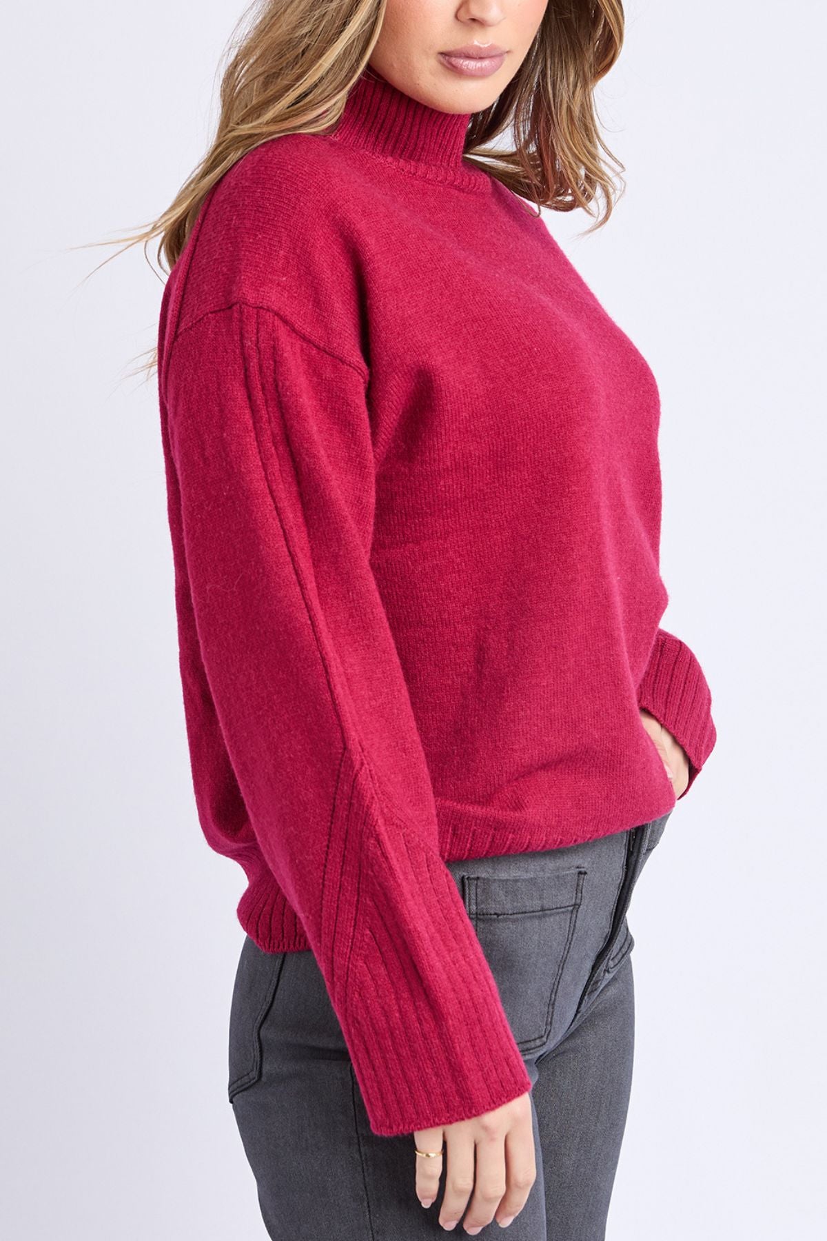 Woman in a loose-fitting, long-sleeve Foxwood Stellar Knit in red and dark gray jeans stands sideways with one hand in her pocket against a plain light background.