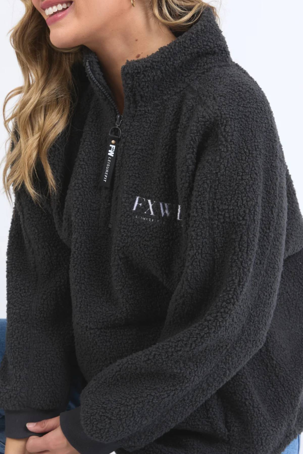 A woman, visible from her lower face and torso, smiles in a black Foxwood Weekend Boucle Crew with "EXW" on the chest. Her long hair drapes over her shoulder, emphasizing the cozy loungewear feel of the fuzzy zip-up pullover.