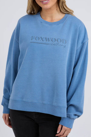 A person wearing the Foxwood Trademark Crew—a loose-fit, light blue unbrushed cotton fleece sweatshirt—paired with black pants against a plain background, makes an ideal addition to any casual wardrobe.
