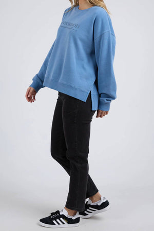 A person stands against a plain background in a loose blue Foxwood Trademark Crew made from unbrushed cotton fleece, paired with black jeans and black Adidas sneakers—an easy, casual outfit. Their head is turned slightly to the side.