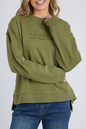 A woman wears the Foxwood Trademark Crew, an olive green unbrushed cotton fleece sweatshirt with ribbed cuffs and a relaxed fit, paired with blue jeans. She smiles warmly—an ideal piece for any casual wardrobe.