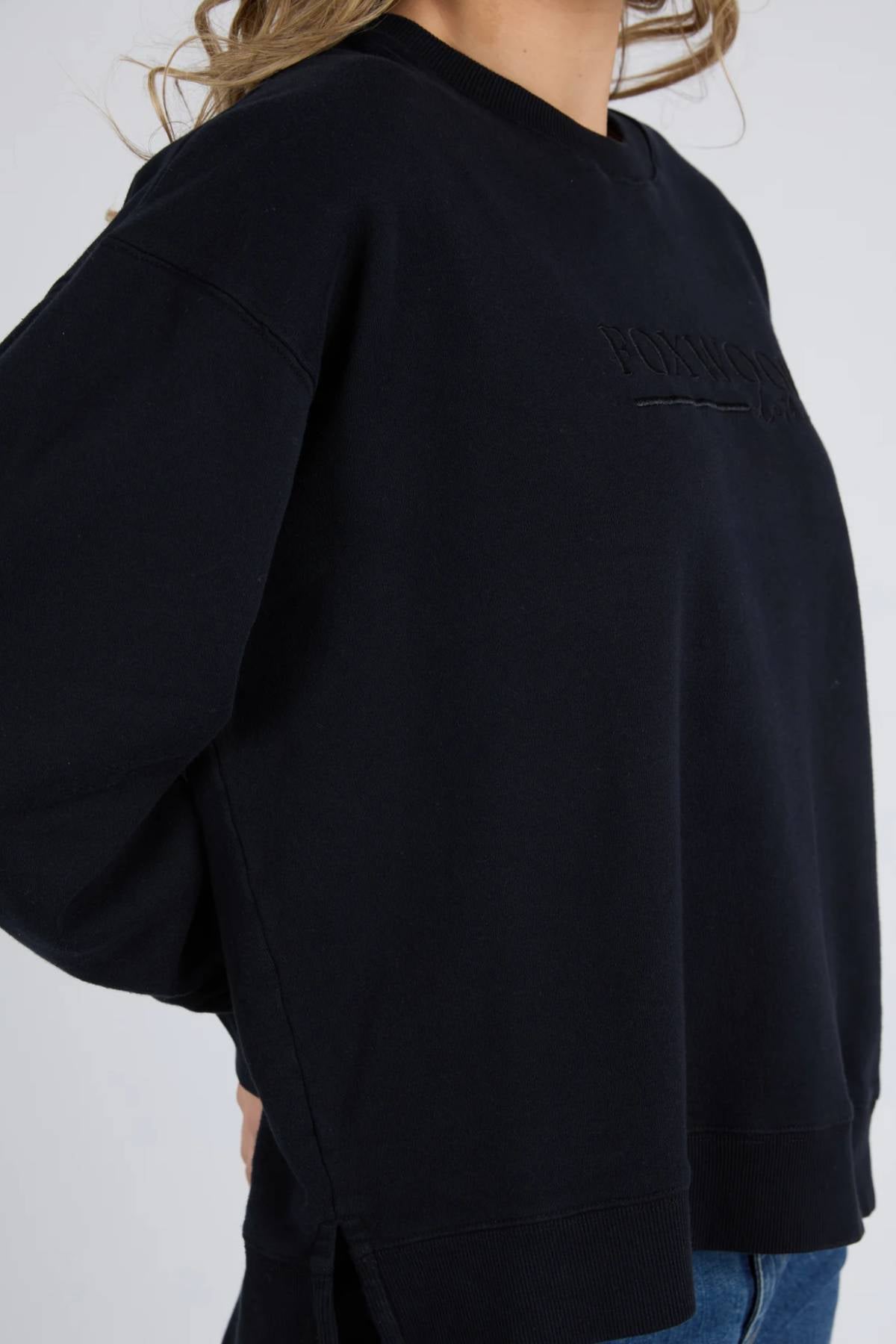 Wearing the loose-fitting Foxwood Trademark Crew in black unbrushed cotton fleece with subtle chest embroidery and blue jeans, a person stands hand on hip. The cropped shot shows their torso and part of their head.