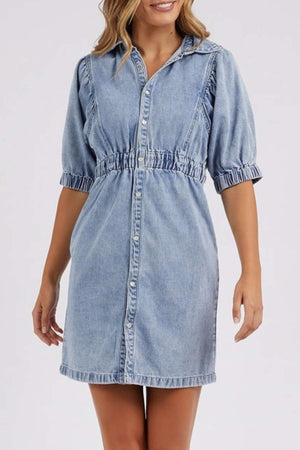 A woman wears the Foxwood Vivian Dress, a light blue stretch denim dress with short puff sleeves, a collared neckline, button-down front, elastic waist, and an above-the-knee length.