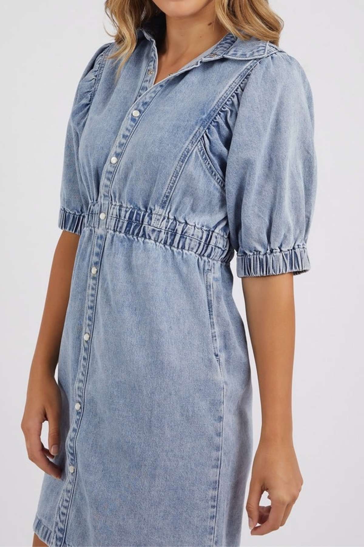 A woman wears the Foxwood Vivian Dress, a light blue stretch denim dress featuring puffed half sleeves, a collared neckline, button-down front, and elastic waist and sleeve details for a chic, comfortable knee-length look.