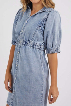 A woman wears the Foxwood Vivian Dress, a light blue stretch denim dress featuring puffed half sleeves, a collared neckline, button-down front, and elastic waist and sleeve details for a chic, comfortable knee-length look.