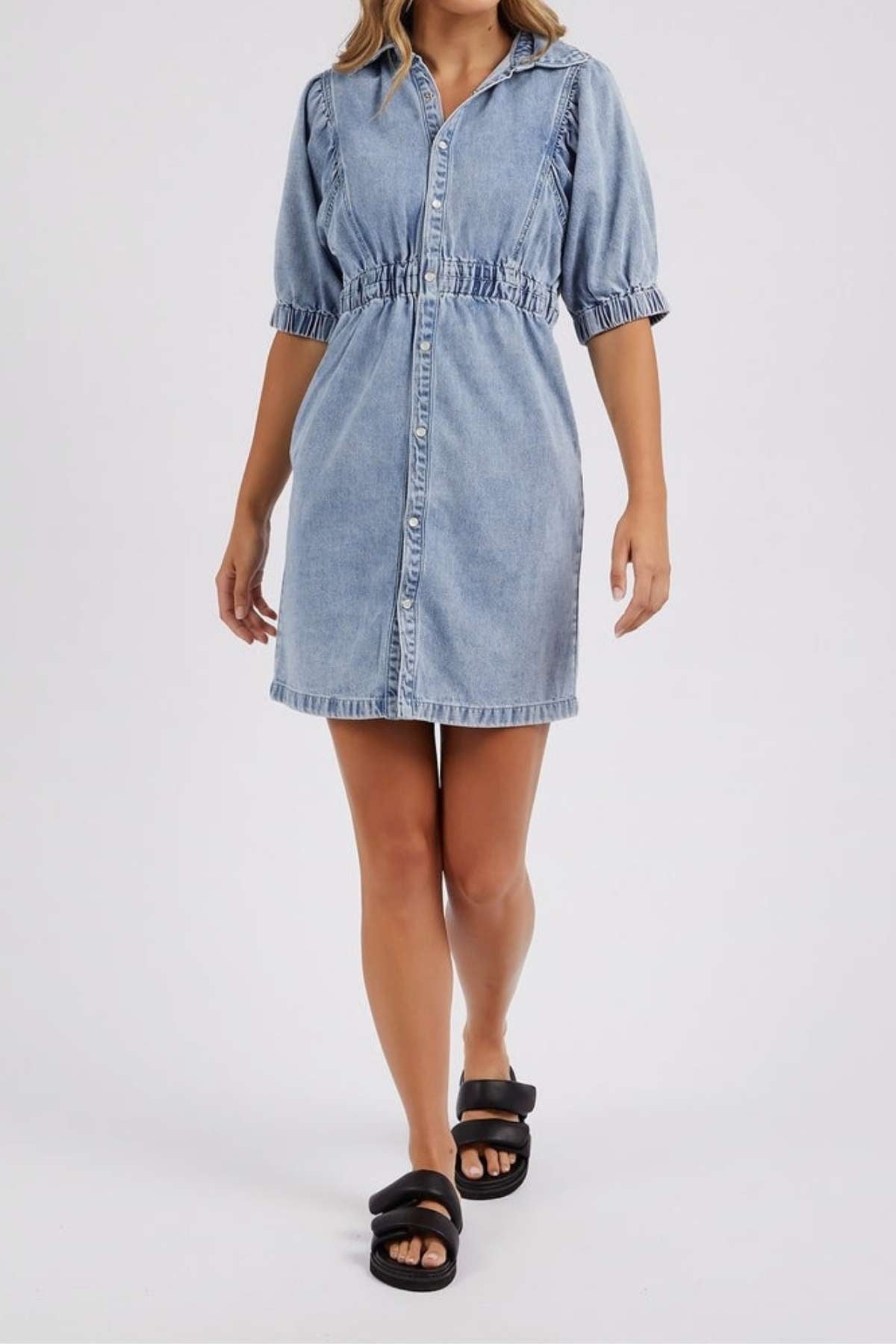 A woman in the Foxwood Vivian Dress—short-sleeve, button-down stretch denim with an elastic waist—pairs it with black slide sandals as she stands before a light background.