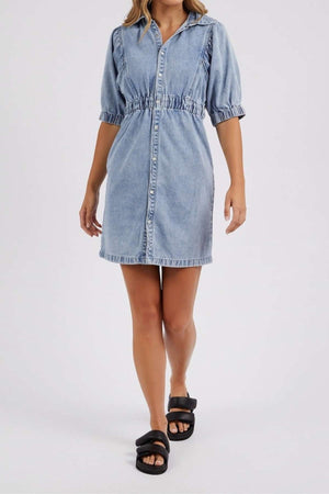A woman in the Foxwood Vivian Dress—short-sleeve, button-down stretch denim with an elastic waist—pairs it with black slide sandals as she stands before a light background.