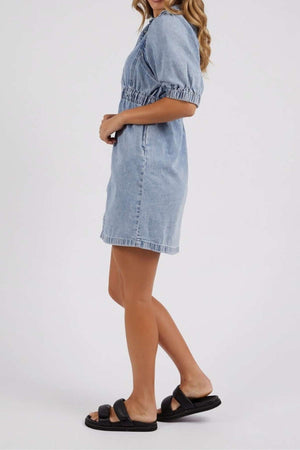 A woman with wavy light brown hair stands sideways in the Foxwood Vivian Dress, a short-sleeved, light blue stretch denim dress with an elastic waist, paired with black slide sandals against a plain white background.