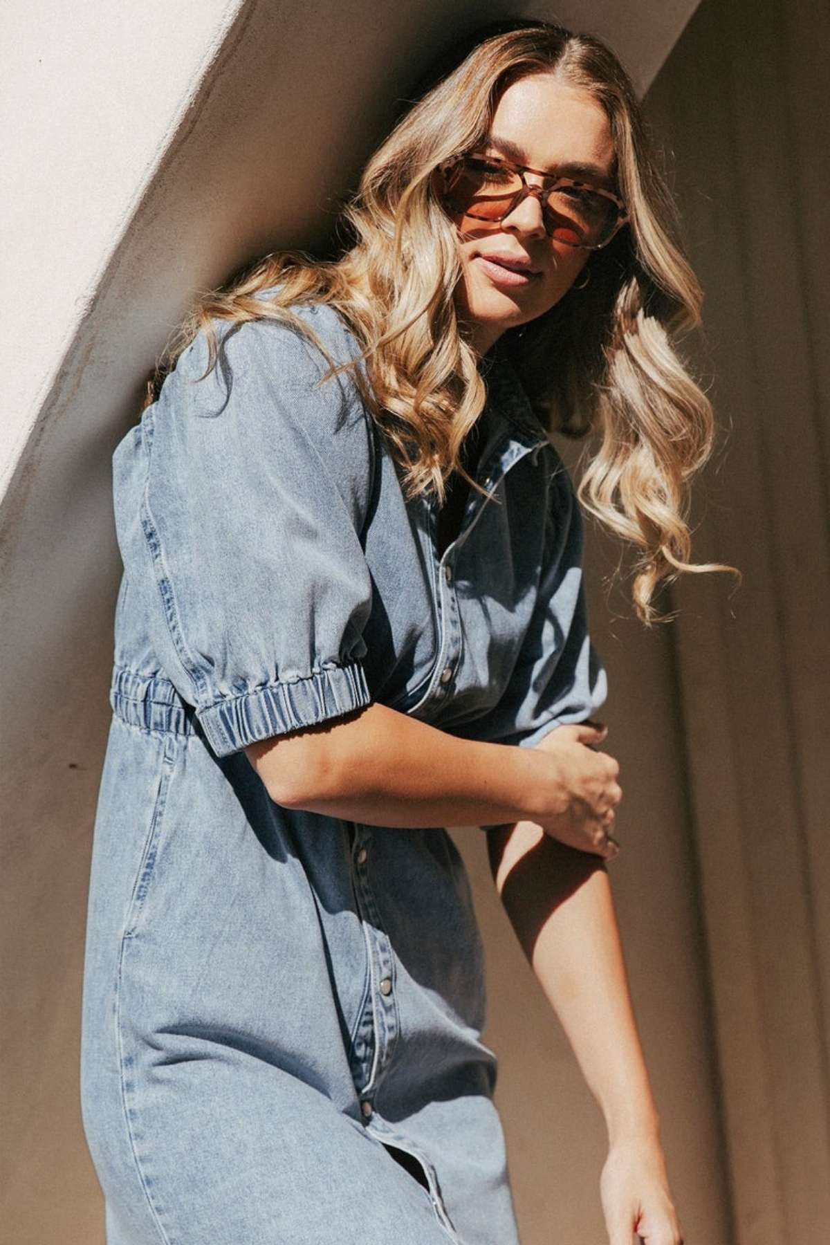 A woman with long wavy blonde hair and sunglasses wears the Foxwood Vivian Dress, a light blue short-sleeve style with an elastic waist, as she stands outdoors in sunlight, leaning slightly against a curved white wall.