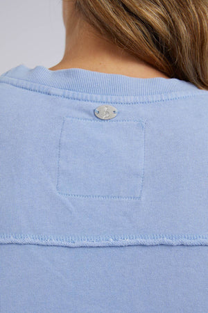 Close-up of a person wearing a comfortable Foxwood Washed Simplified Crew in light blue, made of 100% cotton. The shirt has a visible seam and features a small silver logo near the collar, partially obscured by the person's hair.