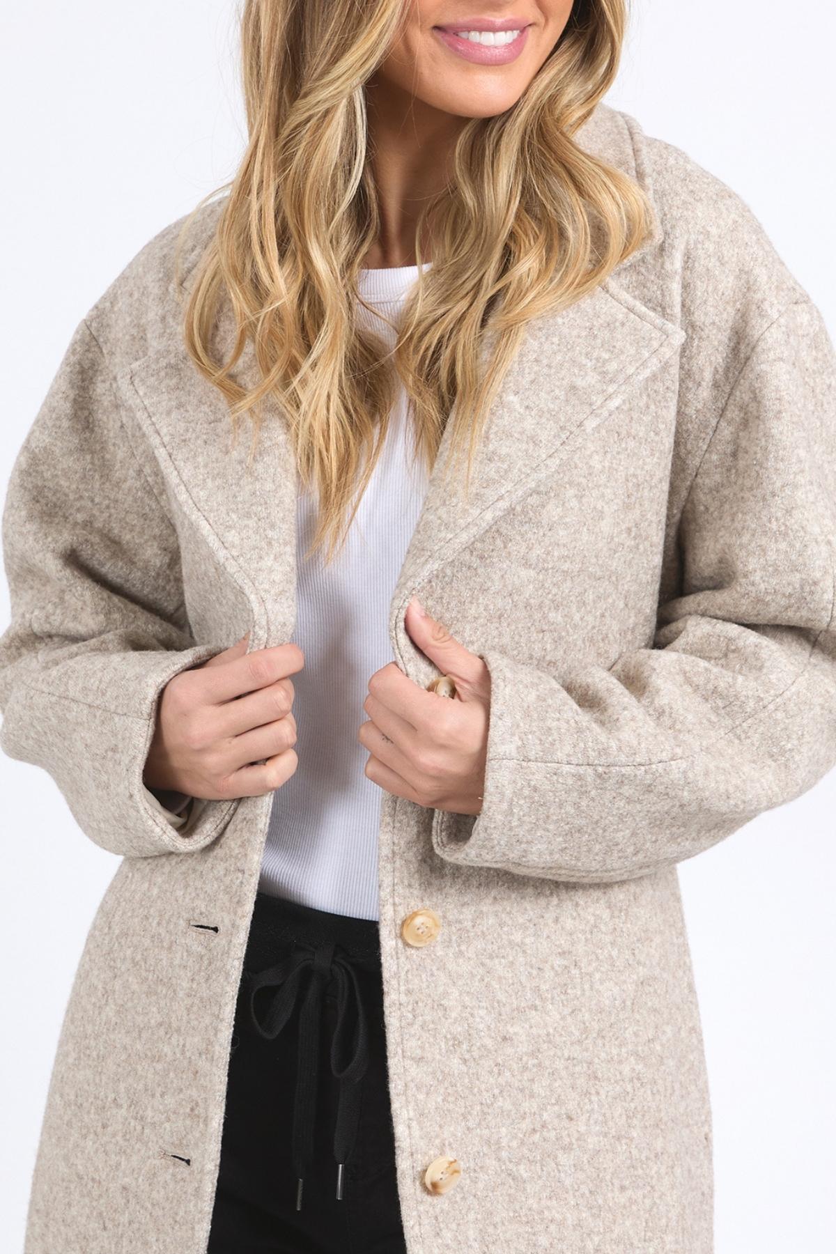 A woman with wavy blonde hair smiles, holding closed her light beige Foxwood Whistler Coat over a white top and black pants. The wool-blend fashion coat is partially concealing her face.