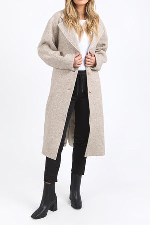 A woman with loose, wavy hair stands against a white background, wearing a long light beige Foxwood Whistler Coat over a white top, black pants, and black ankle boots.
