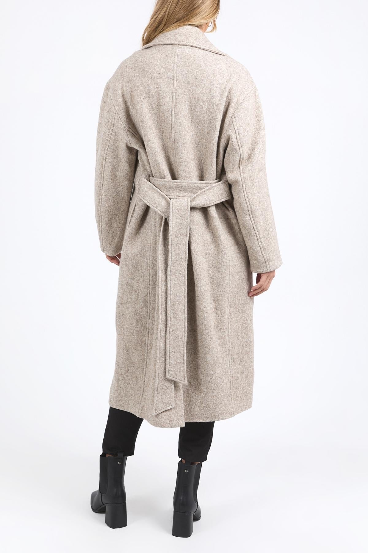 Wearing the Foxwood Whistler Coat—a long, light beige coat—a person in black pants and ankle boots stands with their back to the camera against a plain white background.