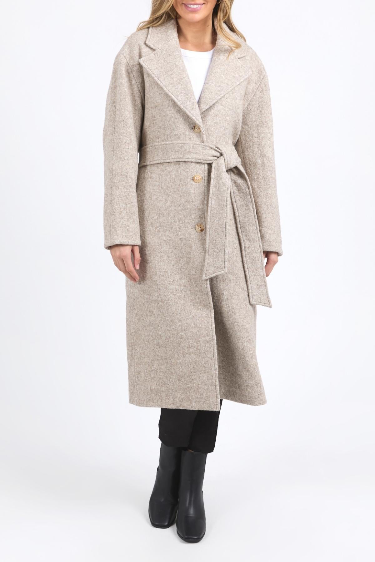 A woman stands against a plain white background wearing the Foxwood Whistler Coat, a long beige coat, styled with black pants and black ankle boots.