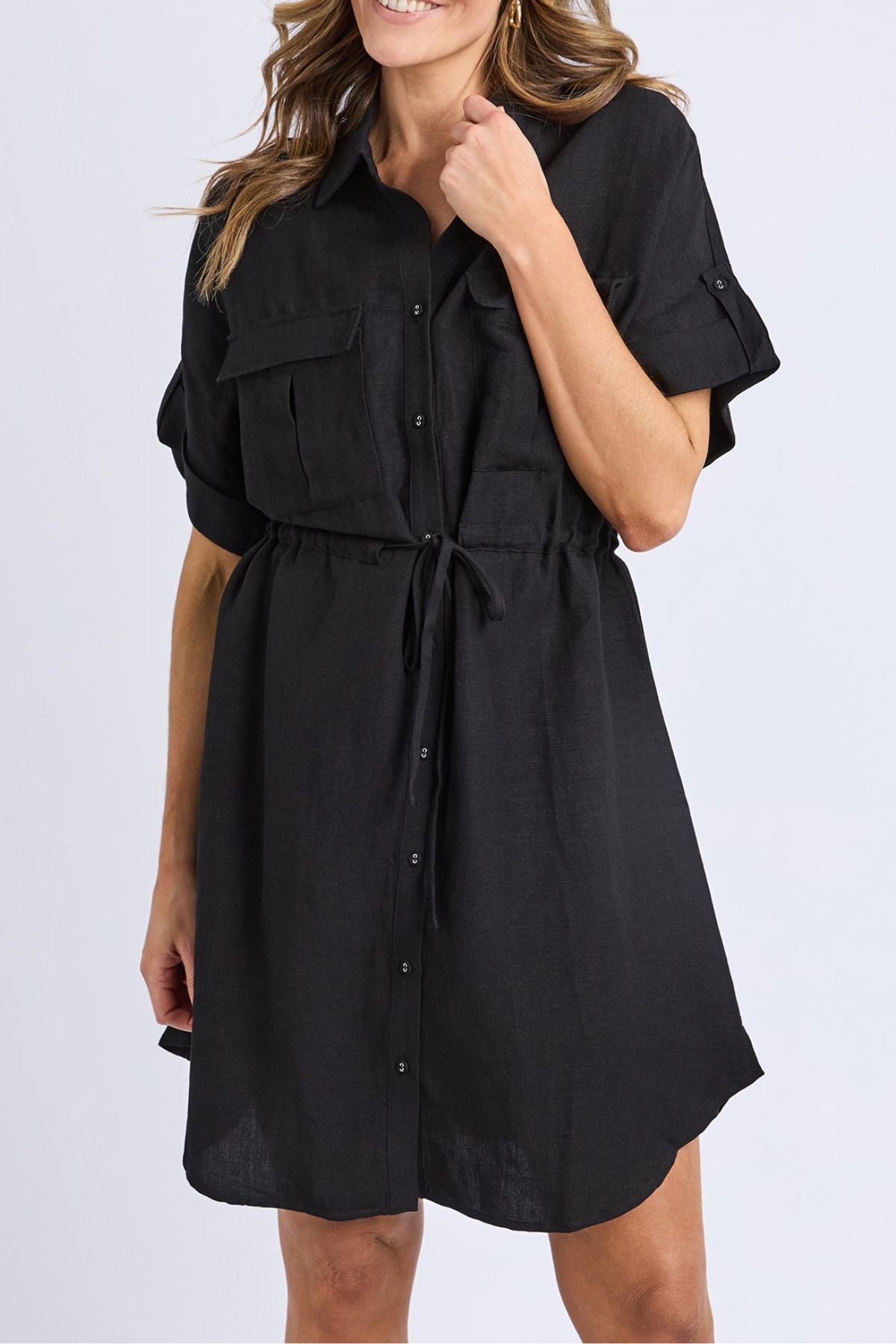 A woman in the Foxwood Willow Shirt Dressโa short-sleeve, button-down linen-viscose dress with a drawstring waist and front pocketsโstands against a plain light background.