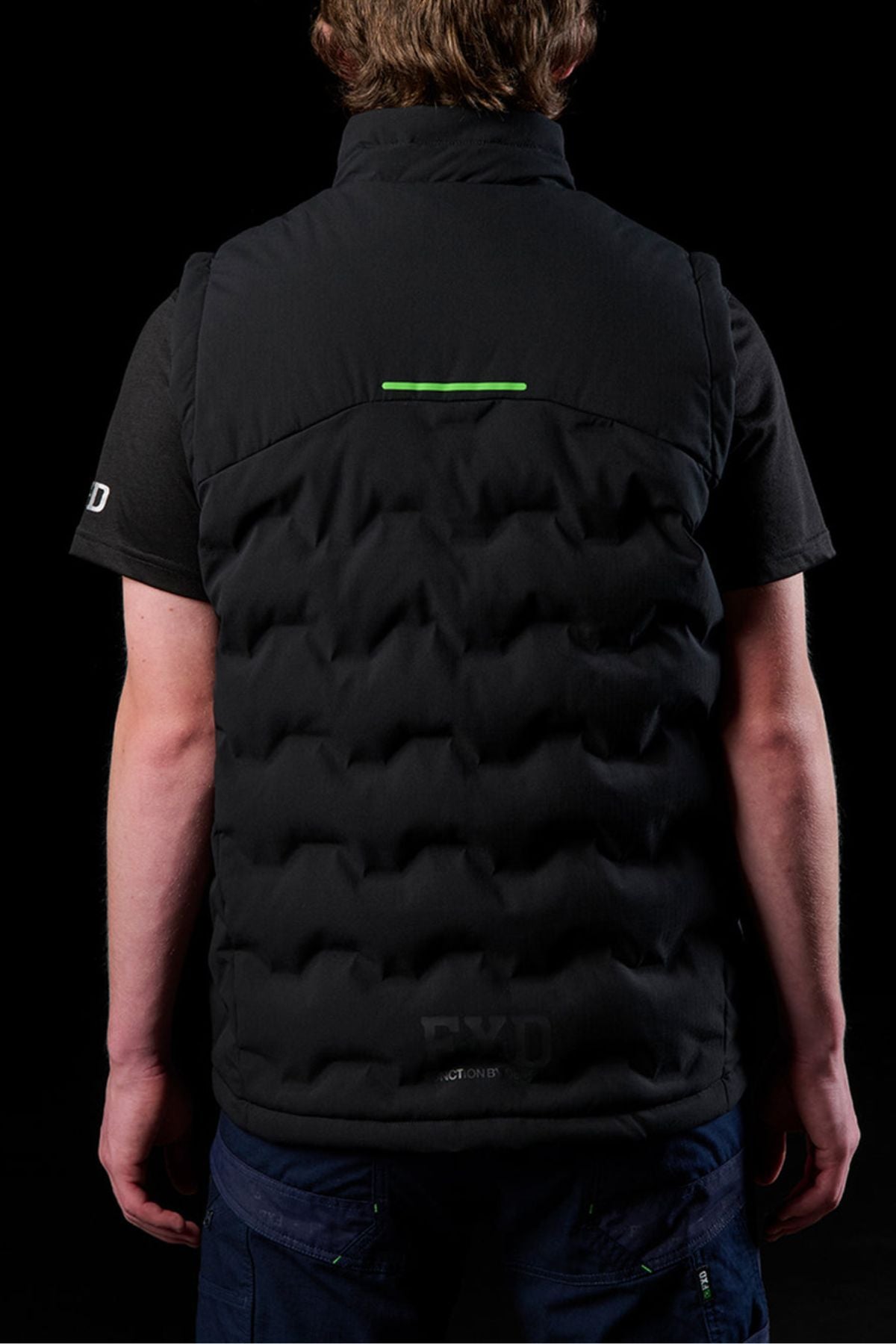 A person is seen from behind wearing the FXD WO-6 Insulated Puffer Vest over a black short-sleeve shirt, set against a black background. The vest features performance fabric and a small green horizontal line near the top.