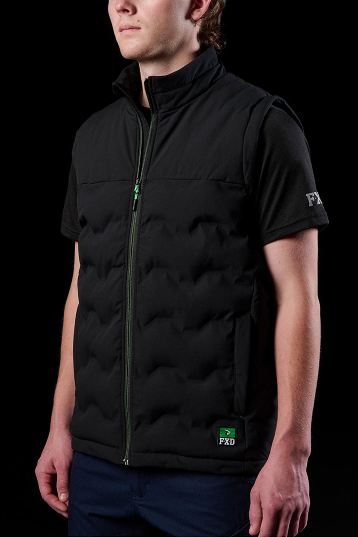 Wearing the FXD WO-6 Insulated Puffer Vest, a black, sleeveless performance vest with green zipper and FXD logo, a person stands against a black background over a matching FXD shirt.