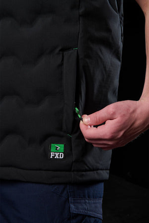 A person pulls the green zipper on the pocket of a black FXD WO-6 Insulated Puffer Vest while wearing navy blue pants. The dark background emphasizes the water repellent vest’s details and prominent FXD logo patch.