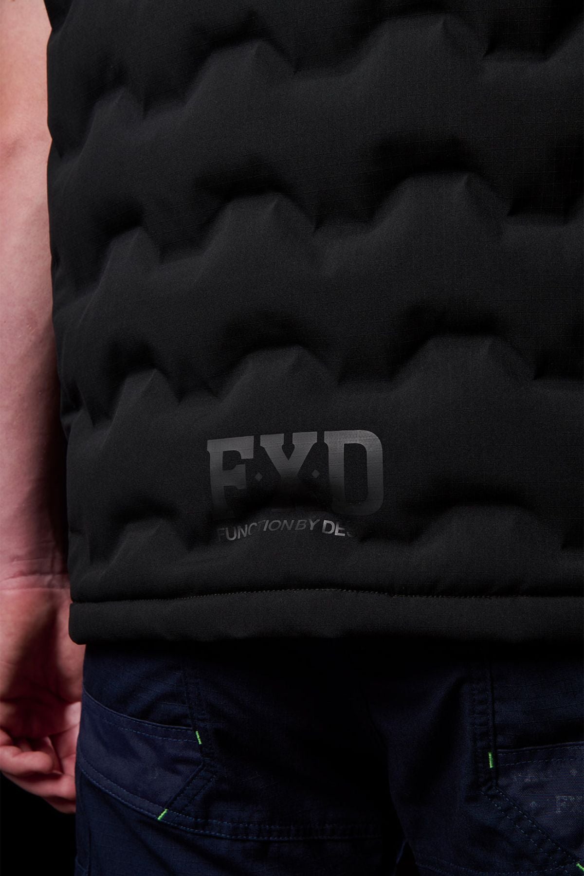 Close-up of a person wearing the FXD WO-6 Insulated Puffer Vest in black with a textured finish and dark blue pants. The FXD logo and "FUNCTION BY DESIGN" are visible near the bottom. Only part of their arm is shown.