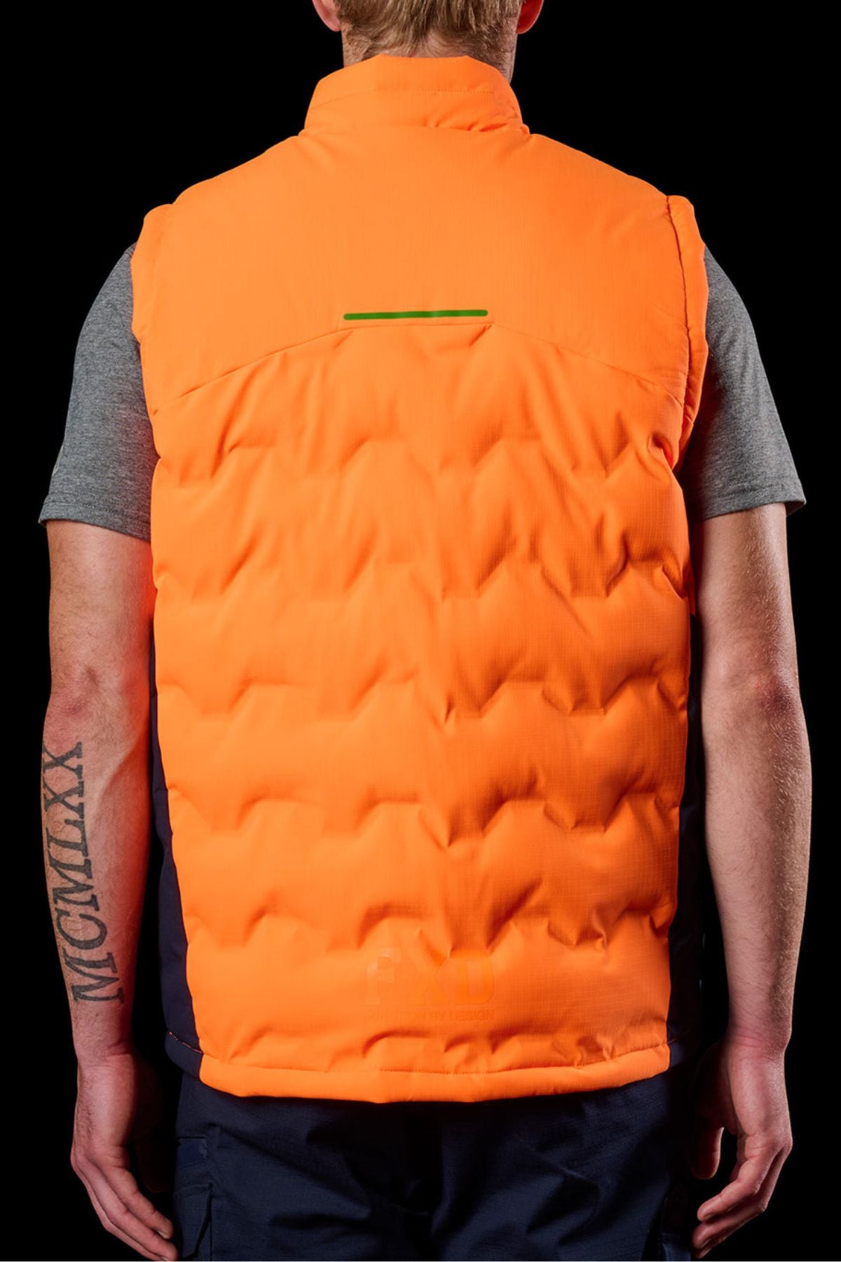 From behind, a light-haired person stands against a black background, wearing the FXD WO-6 Hi Vis Insulated Puffer Vest over a gray short-sleeve shirt, with a Roman numeral tattoo on their right forearm.