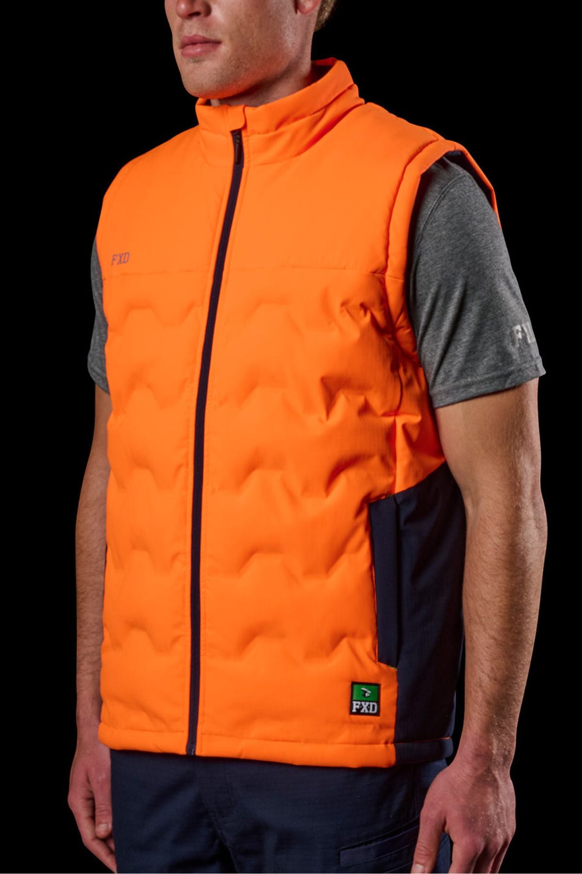 Someone stands against a black background wearing the FXD WO-6 Hi Vis Insulated Puffer Vestβbright orange with navy side panels and the FXD logo on the chest and hemβover a gray short-sleeve shirt.