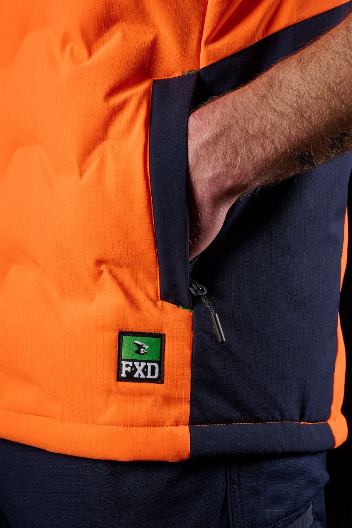 A person wears the FXD WO-6 Hi Vis Insulated Puffer Vest in orange and navy, with a hand in the pocket. The FXD logo patch is visible near the hem, and part of the zipper pull appears next to the pocket on this water-repellent vest.