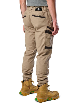A person shown from the back wears FXD WP-12C stretch ripstop work pants with black zipper pockets, a gray shirt, and tan work boots with green soles. The durable utility pants display an "FXD" logo patch on the waistband.