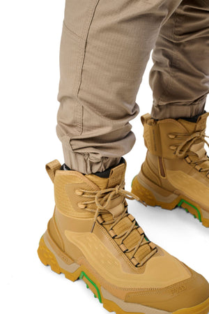 A close-up of a person wearing FXD WP-12C durable utility pants, tucked into tan and yellow high-top hiking boots with green accents, standing on a white background.
