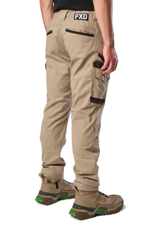 A person models FXD WP-12 tan 2-way stretch ripstop pants with utility pockets and brown details, paired with tan work boots with green soles, standing on a white background facing away from the camera.