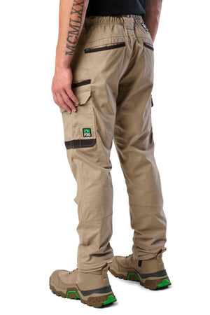 A person wearing FXD WP-12 tan 2-way stretch ripstop pants with black trim, utility pockets, tan work boots with green accents, and a black shirt stands facing away. A tattoo is visible on their left forearm.