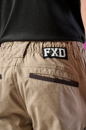 Close-up of a person wearing FXD WP-12 beige durable work pants with utility pockets and a black FXD logo patch above the back pocket, paired with a slightly tucked-in black shirt.