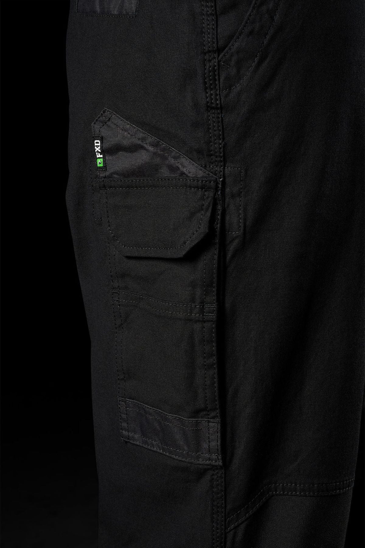 Close-up of black FXD WP-3L Loose Fit Stretch Canvas Work Pants featuring a side pocket with a small green and white label, set against a dark background.