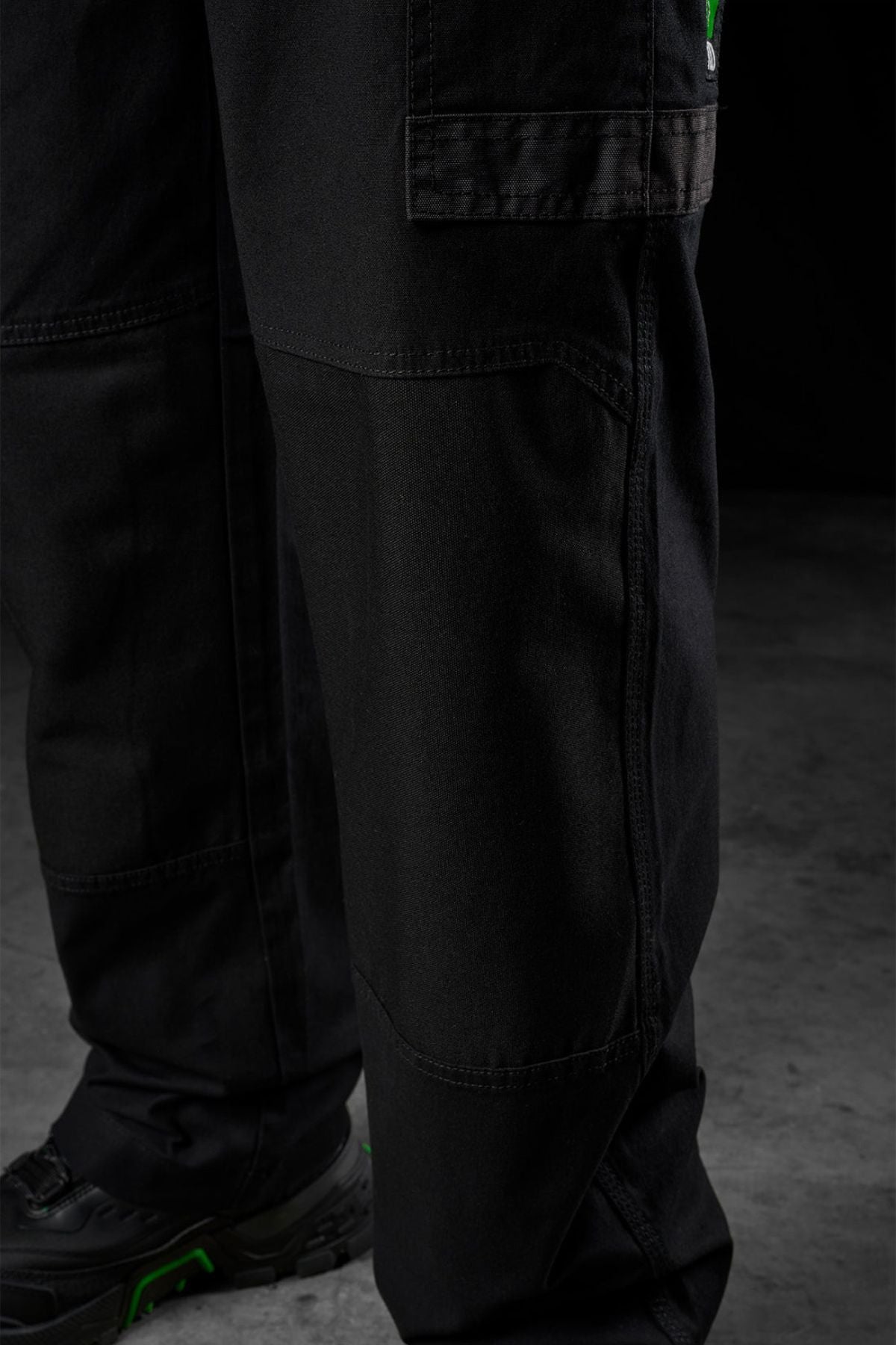 Close-up of a person wearing FXD WP-3L Loose Fit Stretch Canvas Work Pants and black shoes with green accents, standing on a grey concrete floor against a dark background.