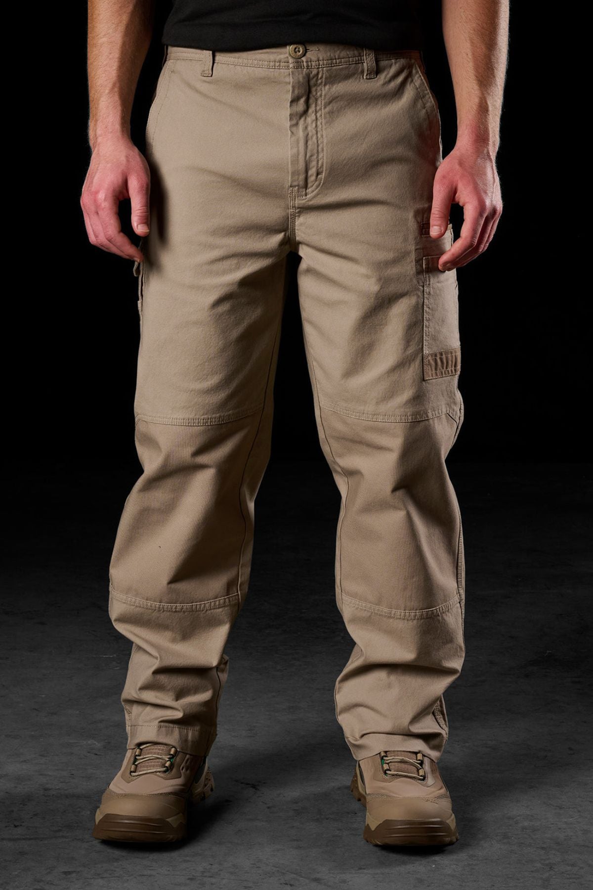 A person models FXD WP-3L Loose Fit Stretch Canvas Work Pants and tan work boots against a dark background, with only the lower half of their body visible and hands relaxed at their sides.