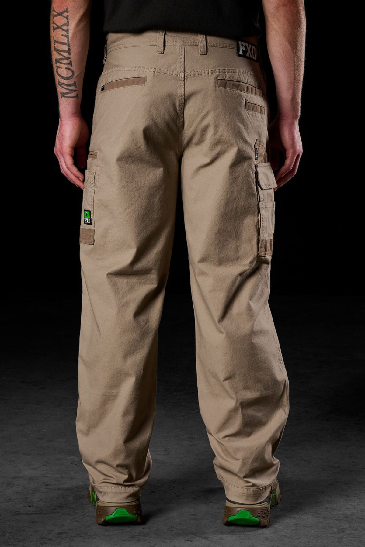 A person faces away, wearing FXD WP-3L Loose Fit Stretch Canvas Work Pants with multiple pockets and green-accented brown shoes. A Roman numeral tattoo is visible on their left forearm against a dark background.