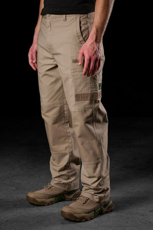 A person wearing tan FXD WP-3L Loose Fit Stretch Canvas Work Pants, a black shirt, and brown work shoes stands against a dark background. The pants offer multiple pockets and reinforced stitching for durability.