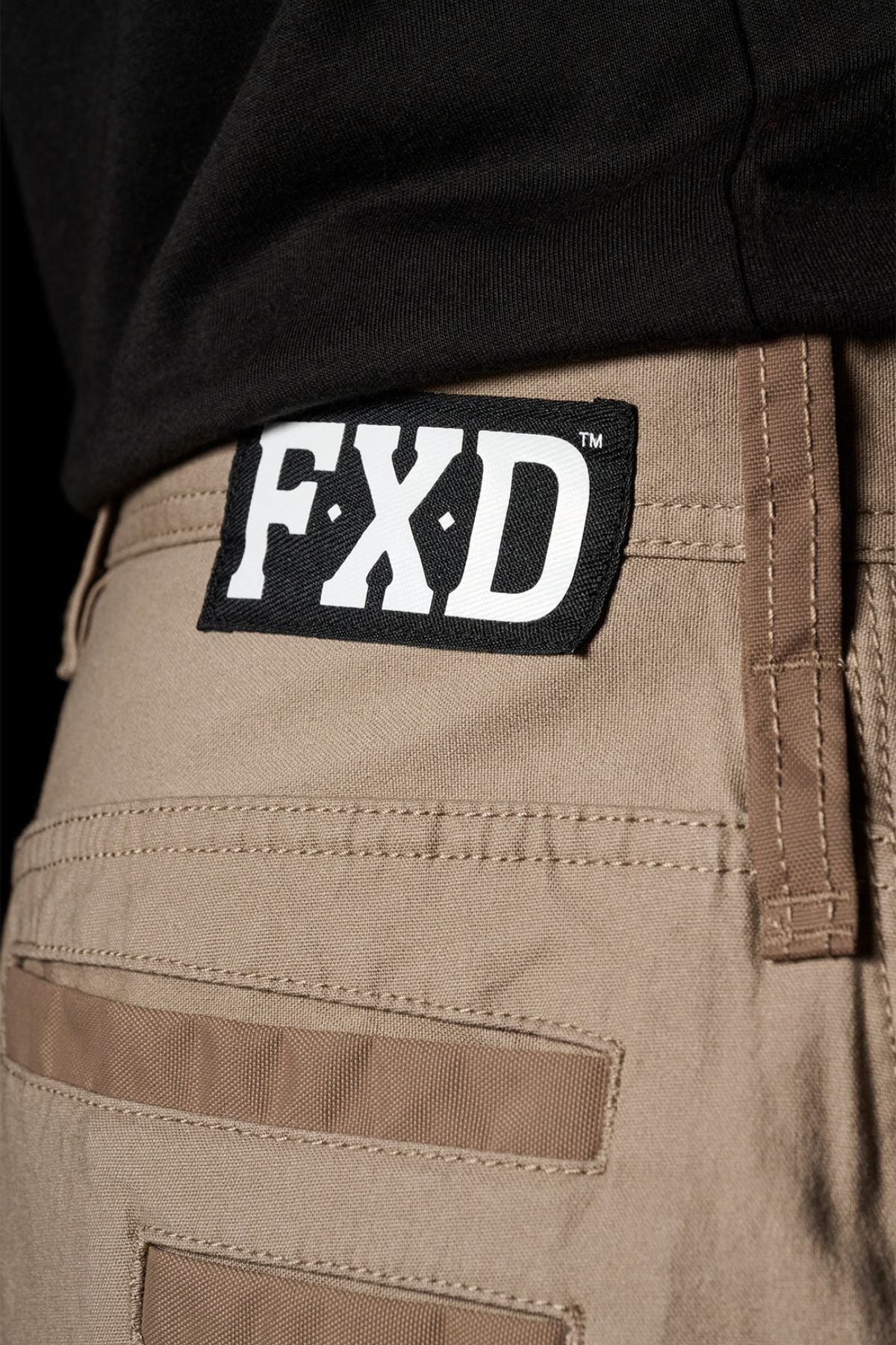 Close-up of FXD WP-3L Loose Fit Stretch Canvas Work Pants in tan, featuring a black and white FXD label on the waistband. They’re styled with a tucked-in black shirt, highlighting their durability and comfortable fit.